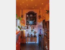 Ein gemütlicher Raum mit orangefarbenen Wänden, einer Wandgestaltung aus geschwungenen Linien und einem hölzernen Regal im Vintage-Stil, das Pflanzen, Keramik und Dekorationsobjekte enthält. Im Vordergrund stehen ein Beistelltisch mit weiteren Dekorstücken und ein Stuhl; an der Decke sind Lichtpunkte verteilt, die einen warmen Eindruck erzeugen.