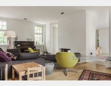 Modernes Wohnzimmer mit Holzboden, einem großen grauen Sofa, einem grünen Designerstuhl und einem runden Couchtisch. Im Hintergrund befinden sich ein Klavier, zwei Sprossenfenster und ein offener Essbereich mit hellen Stühlen.