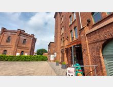 Backsteingebäude in einem Hof mit Pflastersteinen, mit einem runden und einem eckigen Bau im historischen Stil. Im Vordergrund stehen bunte Kinderspielzeuge und Blumentöpfe, rechts ist ein Eingang mit Glas- und Metalltür erkennbar; im Hintergrund ist eine Hecke und blauer Himmel mit Wolken zu sehen.