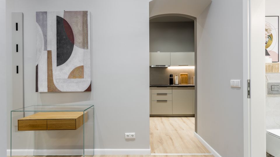 Moderner Wohnungsflur mit hellem Holzboden, einer Glas-Konsole mit Holzschubladen und abstraktem Wandbild. Ein Rundbogen-Durchgang führt in eine kompakte, graue Einbauküche; rechts ist ein offener Türrahmen zu einem Badezimmer erkennbar.