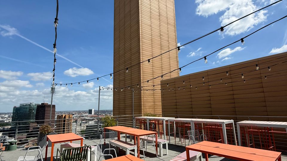 Rooftop terrace with orange tables, mixed seating, and string lights overhead, bordered by metal railings and a wood-paneled wall. A tall tower with tan cladding and a green glass top rises behind the patio, with city skyline views under a blue sky.