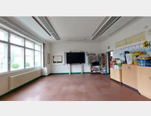 Klassenzimmer mit großen Fenstern an der linken Wand, die viel Tageslicht hereinlassen. Im Raum befinden sich eine digitale Tafel vorne, Regale voller Schulmaterialien an der rechten Wand und eine lange Heizkörperleiste unter den Fenstern.