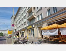 Mehrstöckiges Wohn- und Geschäftsgebäude mit hellbeiger Fassade, großen Fenstern und kleinen Balkonen, darunter Geschäfte und ein Café mit gelben Schirmen und Sitzplätzen im Freien. Im Vordergrund stehen Fahrräder, und ein Verkehrsschild sowie eine Fahrradspur sind sichtbar.