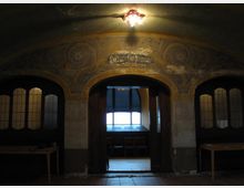 Ein historischer Innenraum mit gewölbter Decke und Verzierungen, die florale Muster zeigen. In der Mitte befindet sich eine offene Doppeltür, die in einen beleuchteten Raum mit Fenstern und Stühlen führt; links und rechts daneben sind Türen mit dekorativen Glaselementen.