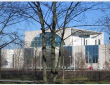 Moderner Regierungsbau mit großen Glasfronten und heller Fassade, umgeben von kahlen Bäumen in einer parkähnlichen Umgebung, bei klarem Himmel. Im Vordergrund sind vereinzelt Bäume und Sträucher zu sehen.