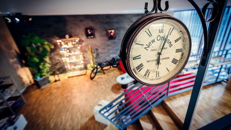 Innenraum im Industrial-Stil mit freitragender Metalltreppe und Holzfußboden; im Vordergrund hängt eine große runde Bahnhofs-Wanduhr. Im Hintergrund sind eine Backsteinwand, dekorative Beleuchtung und ein Fahrrad als Einrichtungselement zu sehen.