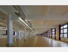 Großer, rechteckiger Innenraum mit hellen Holzböden, weißen Säulenreihen und tiefhängenden Leuchtstofflampen. Der Raum verfügt über große Fenster mit roten Vorhängen, die viel Tageslicht hereinlassen, und wirkt wie eine umgebaute Lagerhalle.