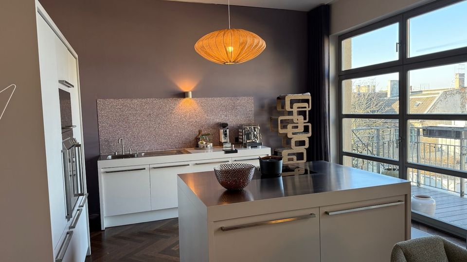 Modern open-plan kitchen with white cabinets, a central island with a dark countertop, and a pendant light above. Large floor-to-ceiling windows and sliding door open to a balcony, bringing in daylight and showing nearby rooftops.