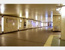 Unterirdischer Tunnel mit cremefarbenen Fliesenwänden, beleuchtet durch vertikale Lichtleisten. An den Wänden befinden sich Wegweisschilder, ein Münztelefon und Poster, während am Ende des Tunnels Glastüren sichtbar sind.