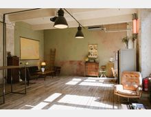 Industriell eingerichteter Raum mit großen Hängelampen, Holzboden und pastellgrünen Wänden. Möbel im Retro-Stil, darunter eine Ledersessel, ein Sofa, Schränke und ein Tisch, sowie dekorative Elemente wie eine Landkarte, ein Globus und hölzerne Leitern an der Wand.