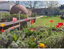 Ein gepflegter Garten mit blühenden roten, gelben und rosa Tulpen, einem Holzsitzbank und einer gepflasterten Weganlage. Im Hintergrund sind ein rundes Gebäude aus Backstein, Büsche, Bäume und eine Terrasse mit Gartenmöbeln zu sehen.
