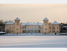 Schloss in sandfarbener Architektur mit zwei symmetrischen Türmen und schneebedecktem Dach, umgeben von einer winterlichen Landschaft; im Vordergrund eine verschneite Fläche und ein zugefrorener See.
