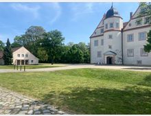 Schlossanlage mit einem historischen Gebäude, das einen weißen Fassadenanstrich mit rosa Fensterrahmen und einen runden Turm mit Spitzdach aufweist, sowie einem Nebengebäude mit rotem Satteldach. Im Vordergrund eine Rasenfläche und gepflasterte Wege, umgeben von Bäumen.