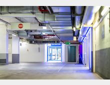 Untergeschoss eines Parkhauses mit weiß gestrichenen Wänden, blauen Hervorhebungen und Beschilderung zu Parkebenen und Kassenautomaten. Im Hintergrund befindet sich ein beleuchteter Durchgang mit Schiebetür und Sicherheitskameras.