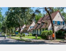 Straßenszene in einem ruhigen Wohngebiet mit Reihenhäusern im Landhausstil, hell verputzt und mit roten Ziegeldächern. Große Bäume säumen die Straße, während zwei Personen auf Fahrrädern entlangfahren; im Vordergrund stehen ein Straßenlampenmast und ein orangefarbener Briefkasten.