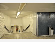 Treppenabgang in einer U-Bahn-Station mit weißen Wandfliesen und blauer Mosaikwand auf der rechten Seite. Im Hintergrund ist der Bahnsteigbereich mit Beschilderung und einem Zugangssperrengerät zu sehen.