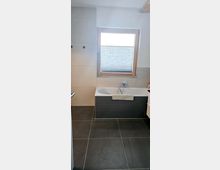 Modernes Badezimmer mit grauen Boden- und Wandfliesen, einer Badewanne mit dunkler Verkleidung und einem Fenster mit halbgeschlossenem Plissee-Rollo. Die Wände sind in hellen Tönen gehalten und die Badewanne ist mit einem Handtuch dekoriert.