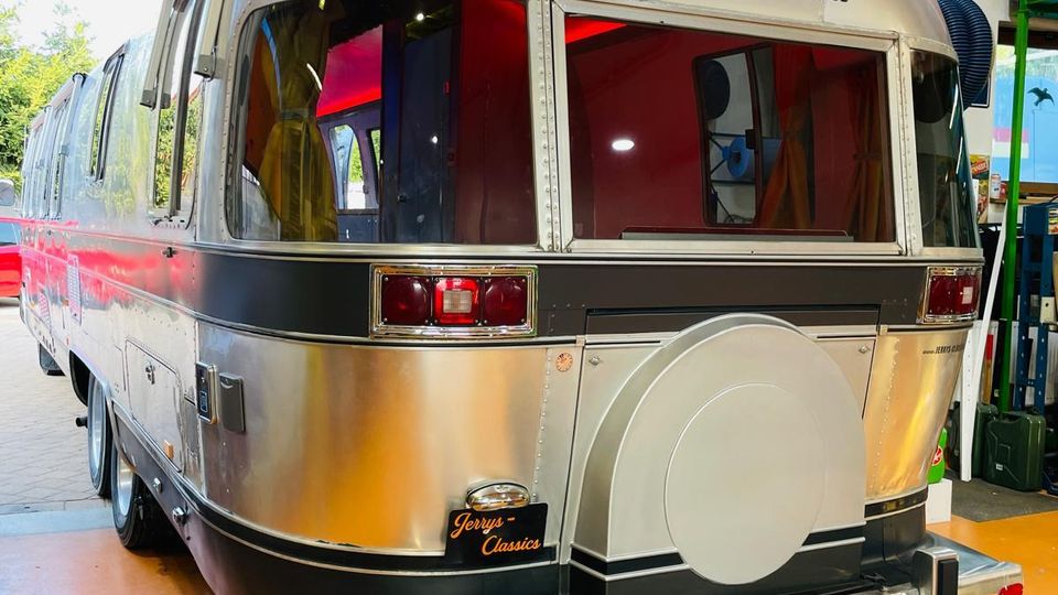 Silberner Airstream-Wohnanhänger von hinten in einer Werkstatt- oder Garagenhalle mit orangefarbenem Rolltor, sichtbaren Leitungen und Regalen. Am Heck sind große Fenster, Rückleuchten, eine Reserveradabdeckung und eine ausklappbare Trittstufe zu sehen.