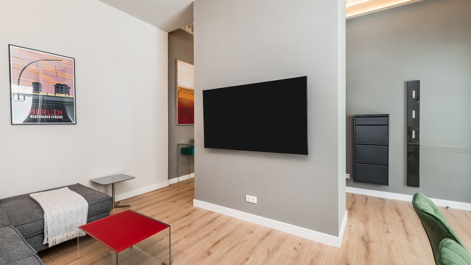 Modernes Wohnzimmer mit hellen Wänden und Holzfußboden, zentraler grauer Trennwand mit wandmontiertem Flachbildfernseher. Links stehen ein graues Ecksofa mit rotem Glastisch und Wandbild, rechts öffnet sich der Blick in einen angrenzenden Bereich mit schwarzen Wandpaneelen und indirekter Deckenbeleuchtung.