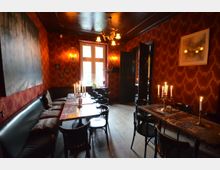 Ein gemütlicher Raum mit dunkelroten, ornamental gemusterten Wänden, schwarzen Holzmöbeln und langen Tischen, die mit Kerzenleuchtern dekoriert sind. Große Fenster lassen Tageslicht herein, während Wandlampen und ein Kronleuchter warmes Licht spenden.