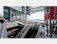 Großer, moderner Bahnhof mit mehreren Ebenen, einer gläsernen Fassade und einem weitläufigen, lichtdurchfluteten Innenraum. Rolltreppen verbinden verschiedene Ebenen, Geschäfte und Werbeplakate flankieren den Bereich, während Reisende zu Fuß oder mit Gepäck unterwegs sind.