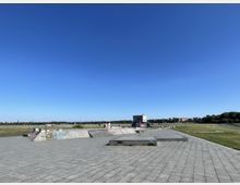 Offener Skatepark auf einer großen, gepflasterten Fläche, umgeben von Rasen und niedrigen Gebäuden im Hintergrund. Der klare blaue Himmel dominiert den oberen Bildbereich.
