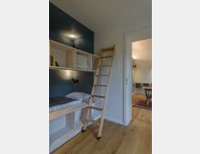 Minimalistisches Schlafzimmer mit eingebautem Einzelbett, Holzregalen und einer Leiter aus hellem Holz. Ein angrenzender Raum ist durch eine geöffnete Tür sichtbar, mit Holzboden, Teppich und modernen Möbeln.