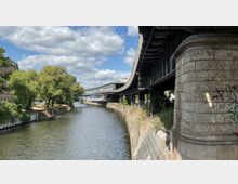 Flusslauf mit Uferböschungen, flankiert von Bäumen links und einer erhöht verlaufenden Stahlbrücke rechts, die ein Bahngleis trägt. Im Hintergrund eine Glas- und Stahlkonstruktion, die als Brücke über den Fluss führt.