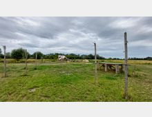 Weite Wiese mit einzelnen Holzpfählen, die im Boden stehen, und einem hölzernen Tisch im Vordergrund. Im Hintergrund sind ein provisorisches Zelt aus Planen sowie Bäume und Buschwerk unter wolkenverhangenem Himmel sichtbar.
