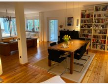 Wohnzimmer mit Essbereich in modernem, warmem Stil. Der Raum hat Holzboden, ein großes Bücherregal, einen Holzesstisch mit sechs Stühlen und eine Sitzecke mit Ledersofas nahe großen Fenstern, die Blick in den Garten bieten.