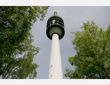 Ansicht eines hohen Fernsehturms mit einer schlanken, weißen Säule und einer runden Beobachtungsplattform, umgeben von grün belaubten Bäumen und grauem Himmel im Hintergrund.