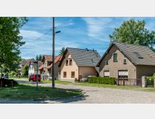 Straßenszene in einer Wohngegend mit Einfamilienhäusern, die überwiegend Satteldächer und helle Fassaden haben. Im Vordergrund verläuft eine gepflasterte Straße, neben der sich ein Wegweiser und eine grüne Rasenfläche befinden, umgeben von Bäumen und kleinen Vorgärten.