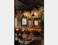 Innenraum eines modernen Cafés mit Holzwänden, an denen Pflanzen und Bilder hängen. Holztische und Stühle stehen auf einem Holzboden, beleuchtet durch industrielle Hängeleuchten; große Topfpflanzen ergänzen die natürliche Dekoration.