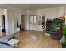 Wohnzimmer mit Holzboden und weißen Wänden, ausgestattet mit einem grünen Kachelofen, einer gepolsterten Sitzgelegenheit, mehreren Zimmerpflanzen und einem kleinen Couchtisch. Im Hintergrund sind zwei Türen, darunter eine Glastür mit Ornamentstruktur, sowie eine schlichte Hängelampe an der Decke.