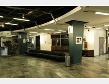 Innenbereich eines öffentlichen Gebäudes mit grauem Marmorboden, einer langen schwarzen Ledersitzbank und modernen Deckenleuchten. Im Hintergrund befinden sich Schilder, ein WC-Hinweis, eine Kasse für ein Planetarium und Dekorationen.