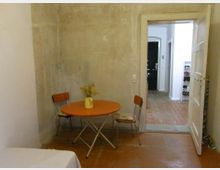 Ein einfacher Raum mit einer unbehandelten Wand, einem orangefarbenen Klapptisch, zwei Stühlen und einer gelben Vase mit getrockneten Gräsern. Im Hintergrund führt eine weiße Tür in einen Flur mit roten Bodenfliesen und offenen Regalen für Schuhe.