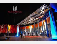 Außenansicht einer Veranstaltungshalle bei Nacht, mit der Aufschrift "Metropolis - Die Halle" oberhalb. Die moderne Glasfassade ist durch orangefarbene Paneele und blaue Beleuchtung akzentuiert, daneben steht eine goldfarbene Skulptur. Im Hintergrund sind rot beleuchtete Felsformationen zu sehen.