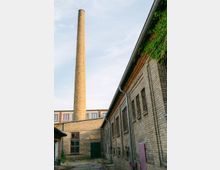 Innenhof eines älteren Industriegebäudes aus hellem Ziegelstein mit hohen Fenstern, Fallrohren und einem mit Efeu bewachsenen Dachrand. Im Hintergrund ragt ein hoher, runder Schornstein aus Ziegelmauerwerk vor einem hellen Himmel auf.