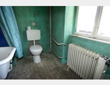 Ein einfacher, abgenutzter Badezimmerraum mit grünen Wänden, einer Toilette, einer alten Badewanne mit Duschvorhang und einem großen Heizkörper unter einem Fenster. Der Holzboden hat sichtbare Gebrauchsspuren.