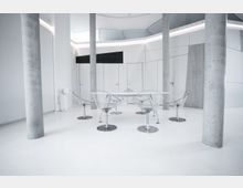 Minimalistischer, moderner Innenraum mit weißen Wänden, glänzendem Boden und Betonstützpfeilern; im Mittelpunkt steht ein Tisch mit transparenten Kunststoffstühlen. Die helle Umgebung wird durch geometrische Wandpaneele und eine indirekte Beleuchtung akzentuiert.