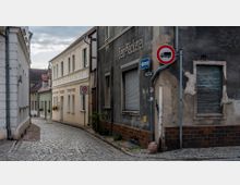 Schmale, gepflasterte Gasse in einer Altstadt mit eng stehenden Wohnhäusern, darunter eine verblasste Fassade mit der Aufschrift „Fein-Bäckerei“. Verkehrszeichen weisen auf eine Tempo-30-Zone und ein Lkw-Durchfahrtsverbot hin.