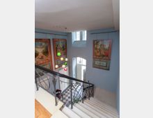 Treppenhaus in einem historischen Gebäude mit weißen Stufen, kunstvollem Metallgeländer und einem Boden aus schwarz-weiß karierten Fliesen. Die Wände sind hellblau gestrichen und mit großformatigen Gemälden geschmückt, während bunte Hängelampen in der Mitte des Raumes angebracht sind.