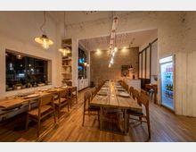 Ein modernes Restaurant mit gemütlicher Atmosphäre, geprägt von langen Holztischen, Holzstühlen und einem Boden aus hellem Holz. Sichtbare Ziegelwände, minimalistische Hängelampen und Details wie Kakteen auf der Fensterbank und ein beleuchteter Kühlschrank prägen den Raum.