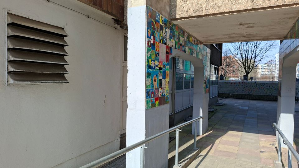 Überdachter Durchgang an einem Gebäude mit weiß verputzten Wänden und Stützen, versehen mit einem bunten Kachelmosaikband entlang der Decke. Ein gepflasterter Weg mit Metallgeländern führt durch den Gang; im Hintergrund sind ein Hofbereich, Bäume und weitere Wohngebäude zu sehen.