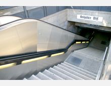 Eingang zu einem U-Bahnhof am Anhalter Bahnhof mit einer breiten Treppe und einer Rolltreppe auf der linken Seite. Über dem Eingang ist ein Schild mit der Aufschrift „Anhalter Bhf.“ angebracht, die Wände sind aus hellgrauem Stein.
