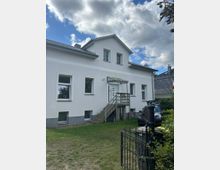 Zweistöckiges Wohnhaus mit weißer Fassade und einem Satteldach, umgeben von einer Rasenfläche. Eine Außentreppe führt zum Eingang, daneben steht ein schwarzes Auto, und im Vordergrund ist ein Metalltor zu sehen.