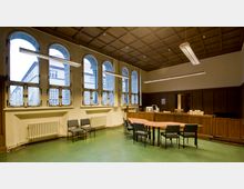 Ein Besprechungsraum mit hohen Rundbogenfenstern, die nach außen blicken und viel Tageslicht hereinlassen. Der Raum hat grüne Böden, eine Holzdecke mit Kassettenmuster sowie dunkle Holzwände; in der Mitte steht ein Tisch mit Stühlen, an der Wand befindet sich ein Heizungskörper.