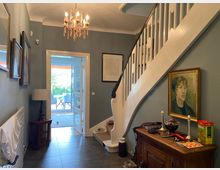 Eingangsbereich eines Wohnhauses mit grauen Wänden, einer weißen Treppe mit Holzgeländer und einem Kronleuchter. Im Vordergrund steht eine antike Kommode mit einer Kerzenhalterdekoration, daneben ein Gemälde, und im Hintergrund öffnet sich eine Tür zu einer Terrasse mit Möbeln.