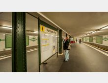 U-Bahn-Station Potsdamer Platz mit einem Fahrgast, der eine Infotafel liest. Die Station hat weiße Kacheln mit grünen Elementen, grüne Stahlträger und einen schwarzen Boden; im Hintergrund sind weitere Passagiere und ein Automat zu sehen.