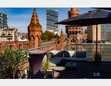 Dachterrasse mit Sitzmöbeln und Pflanzen im Vordergrund, im Hintergrund die Oberbaumbrücke mit ihren charakteristischen roten Backsteintürmen und spitzen Dächern, umgeben von modernen Gebäuden und der Spree.